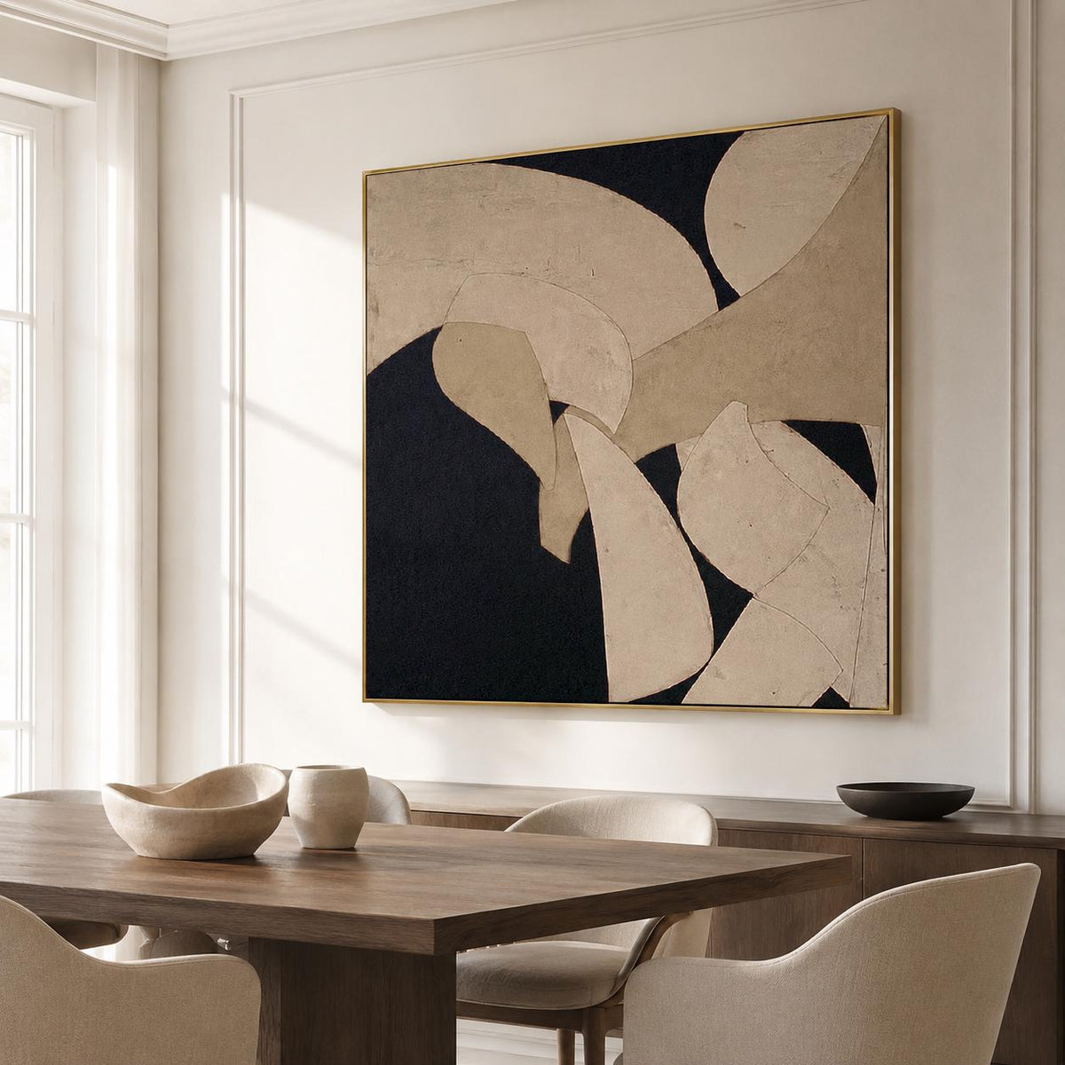 May include: Abstract art piece with a gold frame. The artwork features a composition of beige and black shapes. The painting is displayed in a well-lit room with a wooden dining table and chairs.