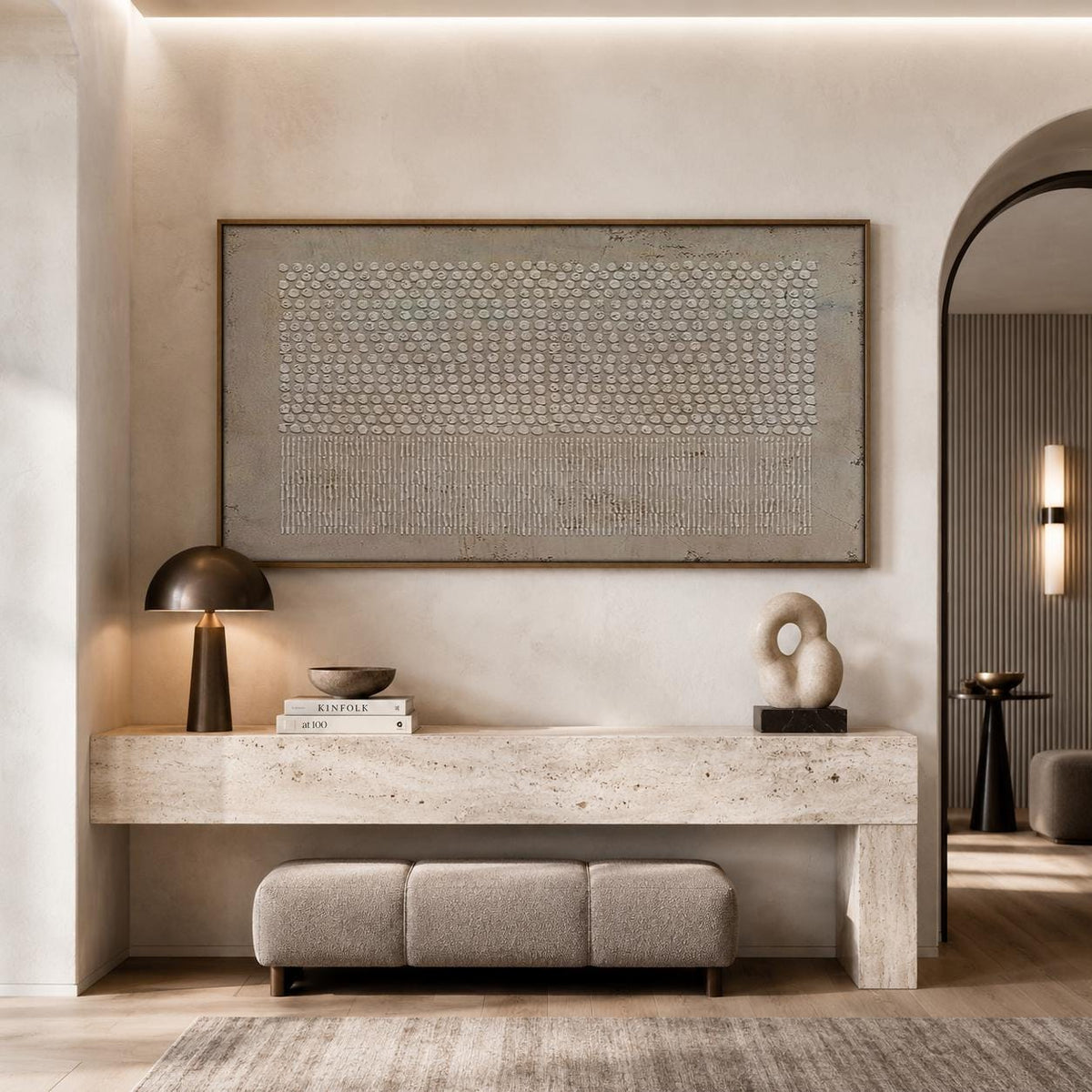 May include: A rectangular wall art piece featuring a textured beige pattern of dots and vertical lines in a thin brown frame, positioned above a long travertine console table with a dome lamp, two stacked books, and a sculptural stone object.