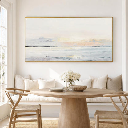 May include: A large rectangular landscape painting with a gold frame. The artwork depicts a serene coastal scene with soft pastel colors of white, blue, and peach. The painting hangs above a wooden dining table.