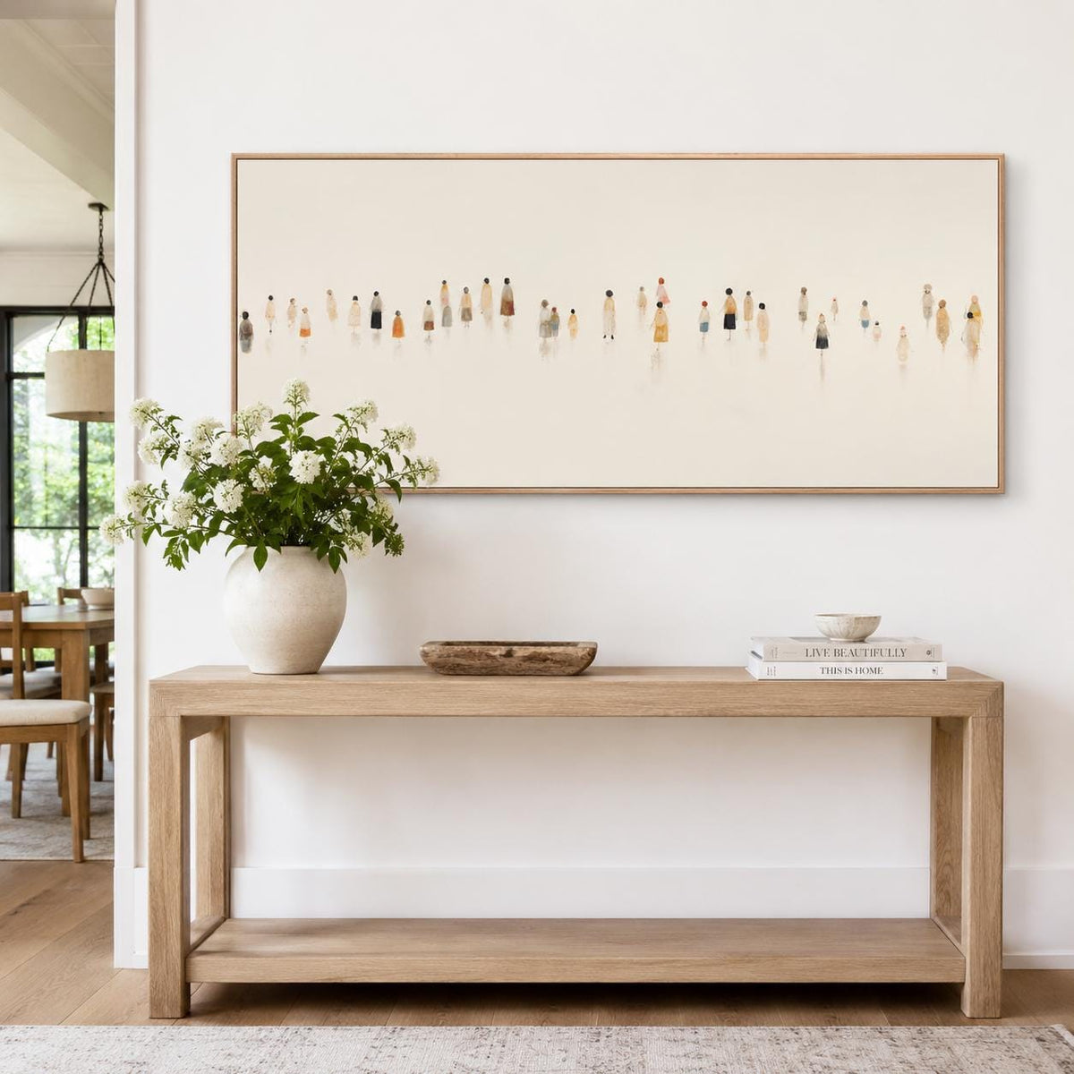 May include: A light wood console table with a rectangular shape and a lower shelf. A large framed artwork hangs above the table, depicting small figures. A white vase with flowers, a wooden bowl, and books are on the table.