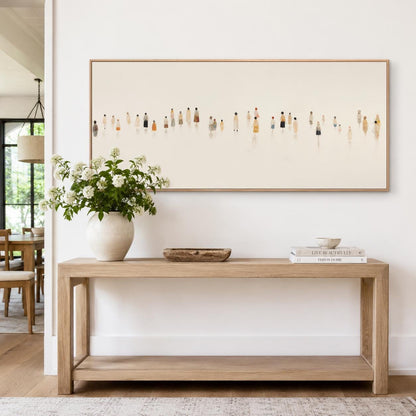 May include: A light wood console table with a rectangular shape and a lower shelf. A large framed artwork hangs above the table, depicting small figures. A white vase with flowers, a wooden bowl, and books are on the table.