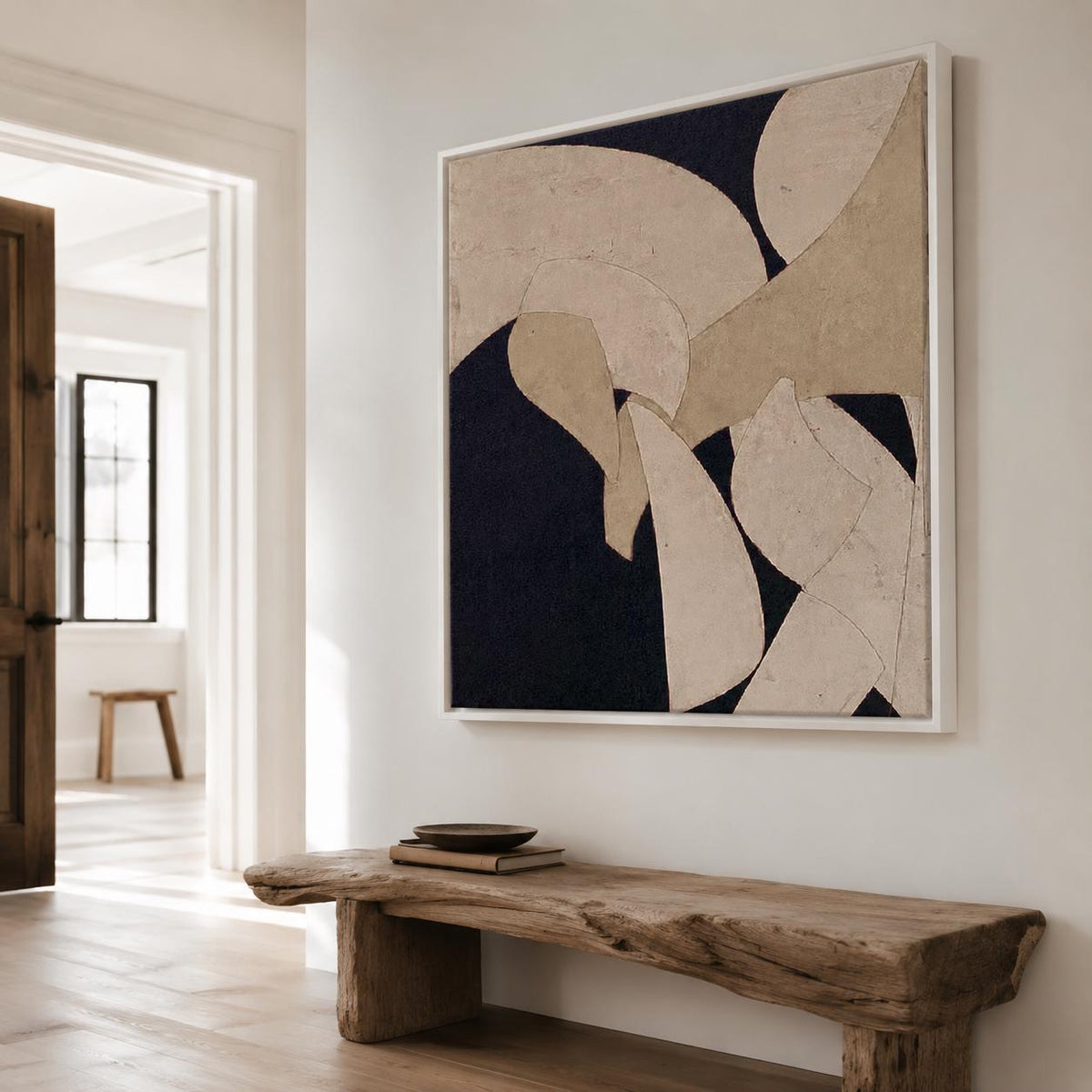 May include: Abstract art piece in a white frame. The artwork features beige and off-white shapes against a dark navy background. The piece hangs on a white wall above a rustic wooden bench.