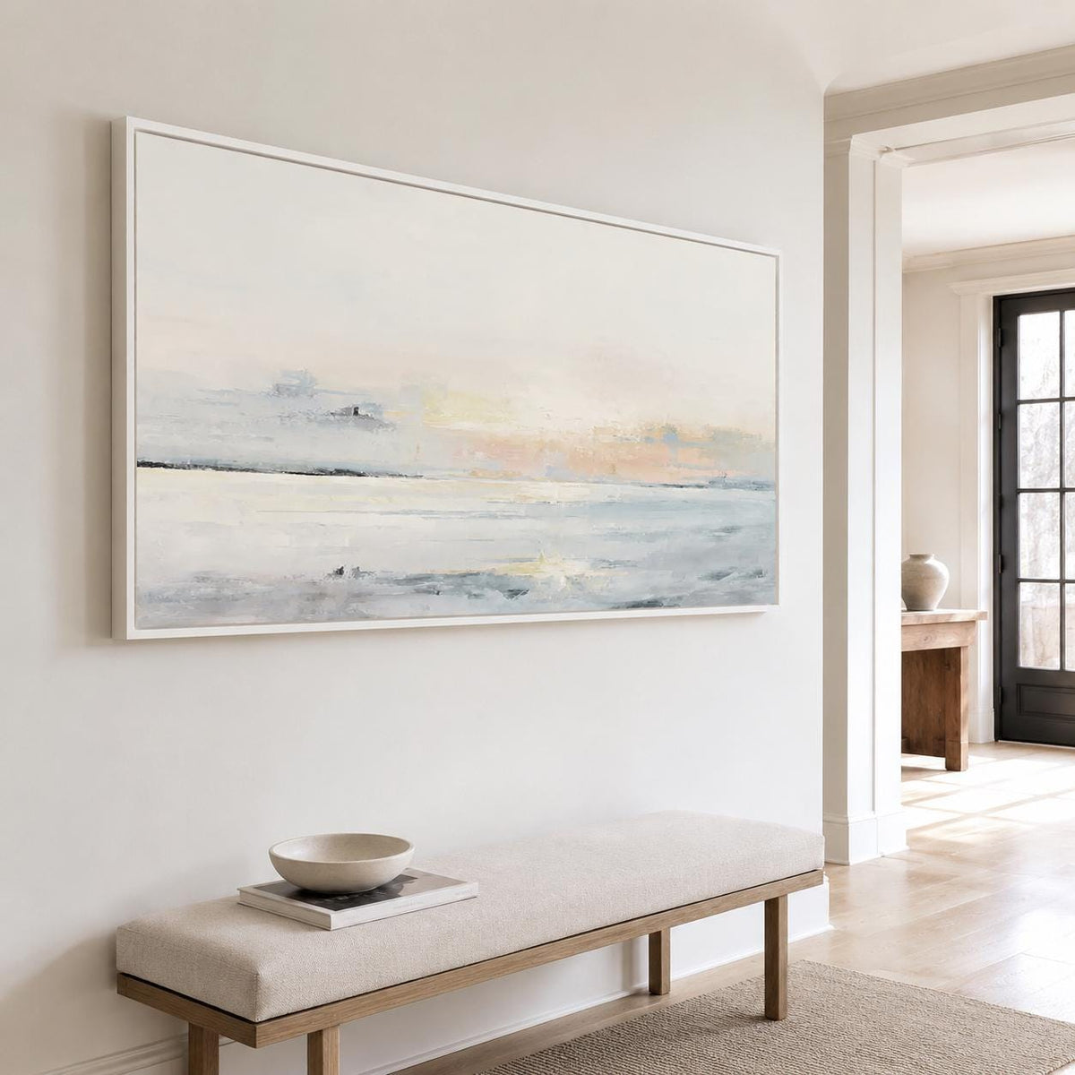 May include: A large rectangular abstract painting in a white frame hangs on a white wall. The artwork depicts a seascape with soft blues, whites, and hints of peach. Below, a beige bench with a wooden frame and a small bowl and book add to the serene interior.