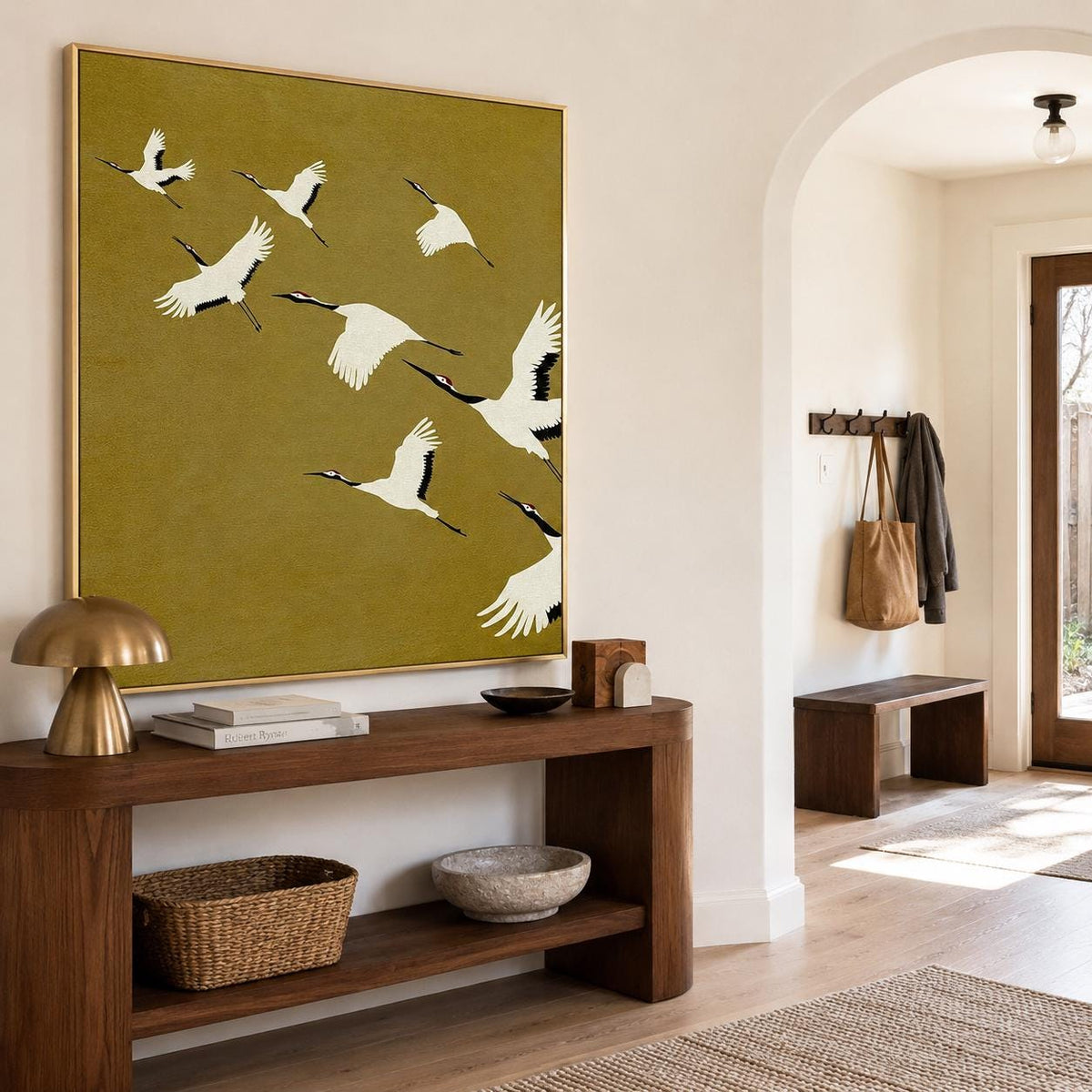 May include: A large square wall art piece featuring a flock of white cranes with black wingtips and head markings flying across a solid olive green background, framed in a thin gold metal border, displayed on a wall above a wooden console table.