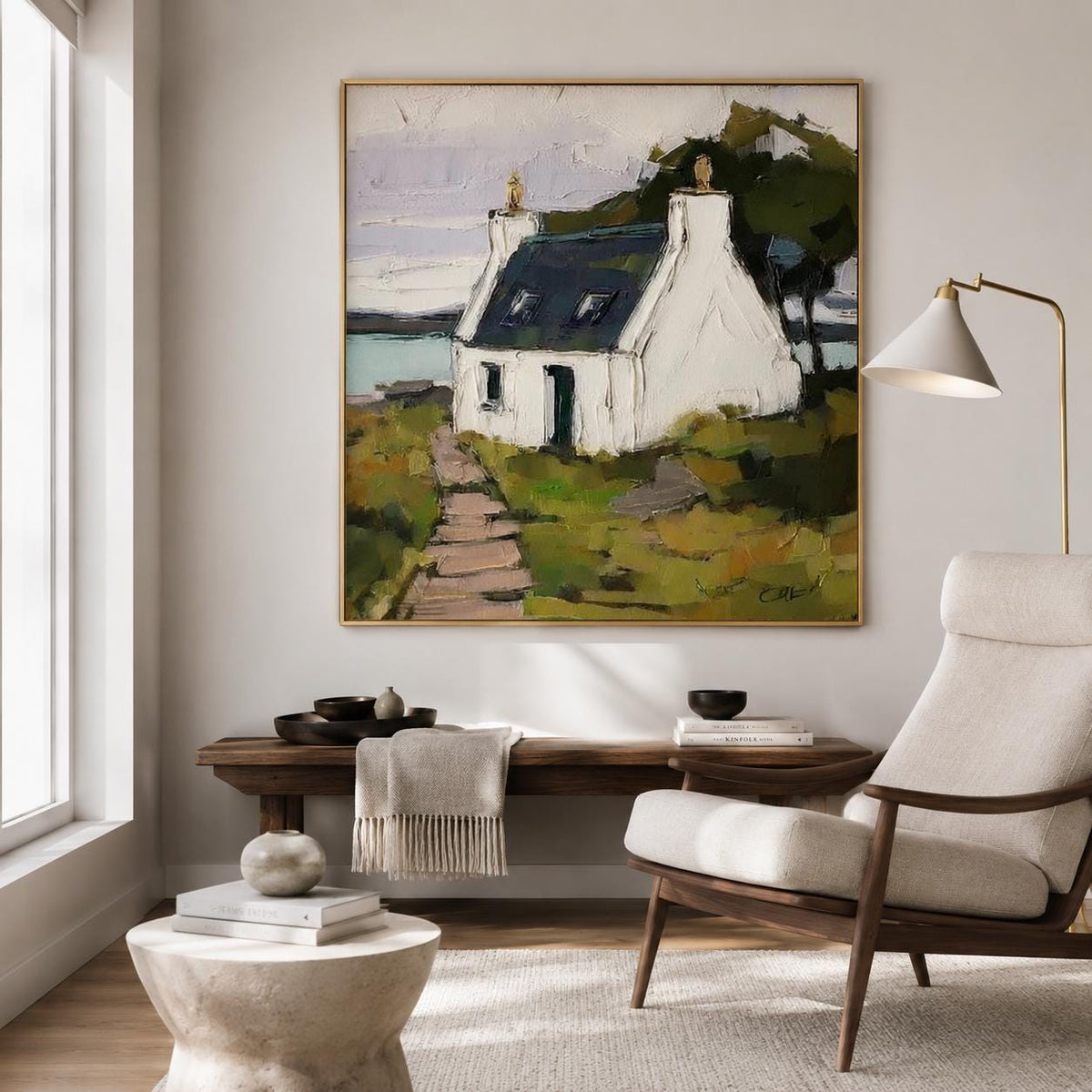 May include: A square oil painting in a gold frame depicting a white cottage with a dark roof, situated on a grassy landscape with a stone path leading to the entrance. The scene includes a body of water in the background and dark green trees.