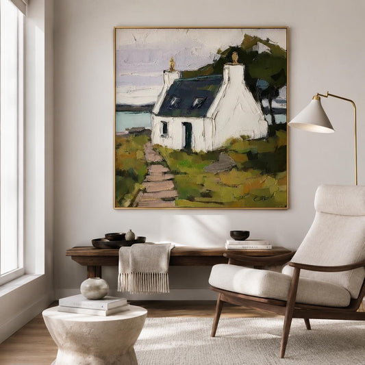 May include: A square oil painting in a gold frame depicting a white cottage with a dark roof, situated on a grassy landscape with a stone path leading to the entrance. The scene includes a body of water in the background and dark green trees.