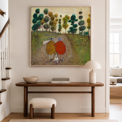 May include: A square painting titled Side by Side featuring two figures in yellow and red coats walking with a small dog on a dirt path through a green landscape with stylized trees. The artwork is framed in light wood and hangs on a wall.