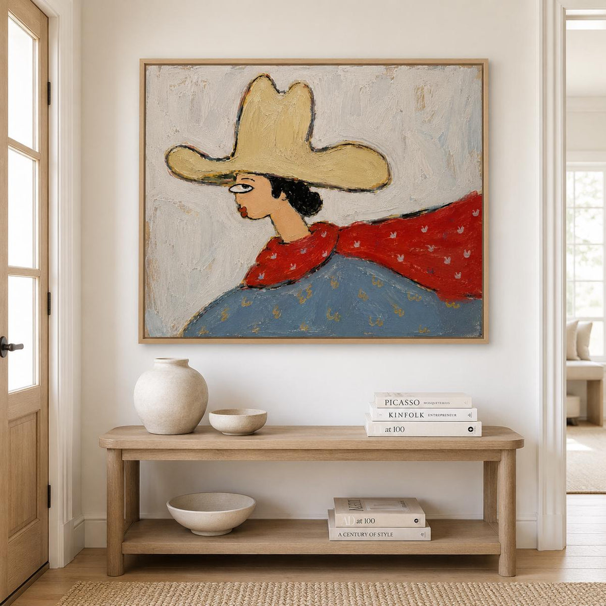May include: A framed painting depicting a figure in a large yellow hat, a red scarf, and a blue garment against a textured off-white background. The artwork is displayed above a wooden console table with a vase, bowls, and stacked books.
