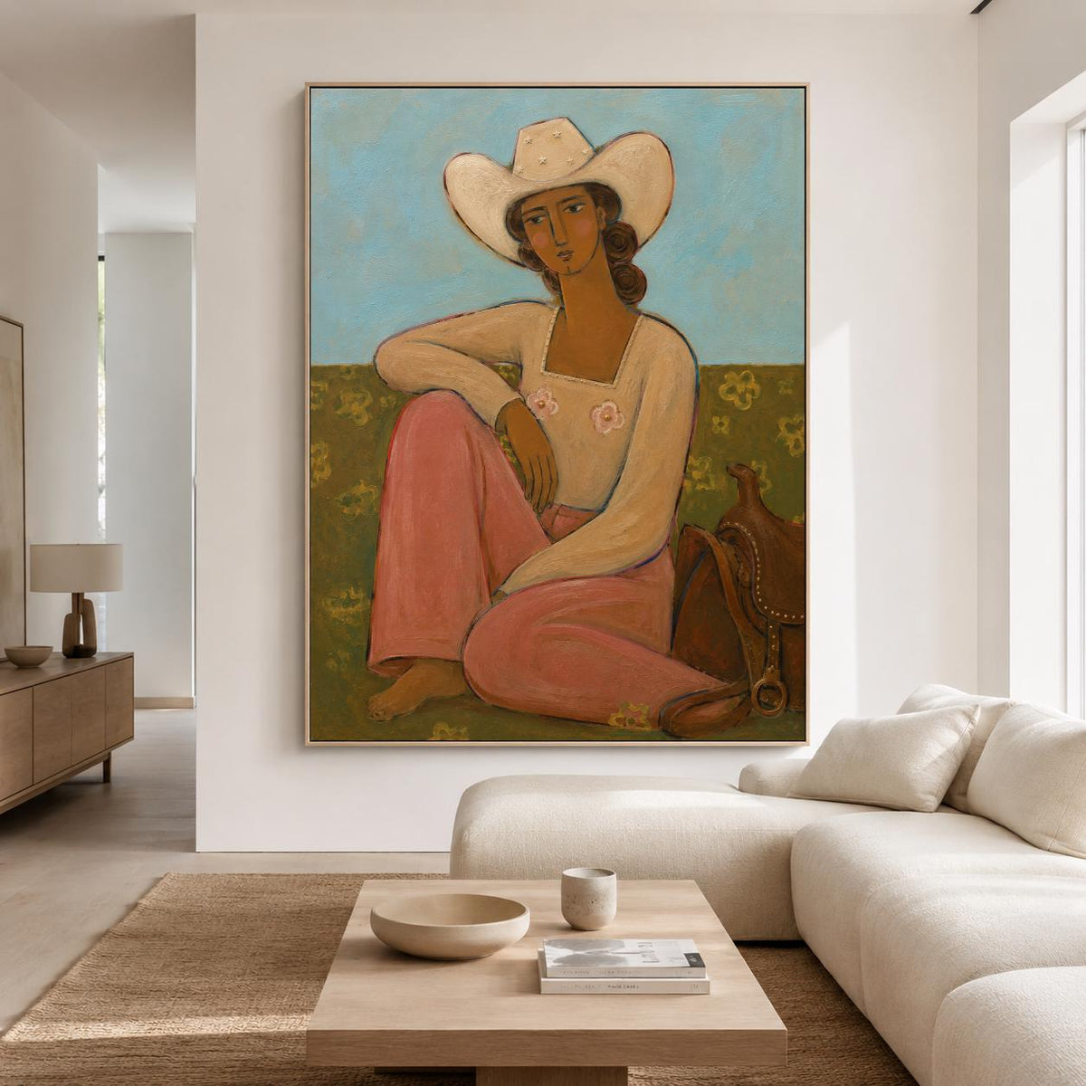 May include: A framed painting depicting a person wearing a white cowboy hat and a long-sleeved cream top with pink floral accents, paired with pink trousers. The figure sits in a field with a brown saddle beside them against a light blue sky.