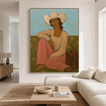 May include: A framed painting depicting a person wearing a white cowboy hat and a long-sleeved cream top with pink floral accents, paired with pink trousers. The figure sits in a field with a brown saddle beside them against a light blue sky.