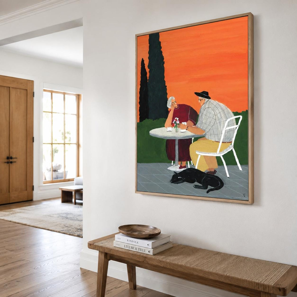 May include: A framed rectangular wall art print depicting two people sitting at a round white table outdoors against a vibrant orange sky. One person wears a dark red dress, the other a plaid shirt and yellow trousers. A black cat lies on the ground.