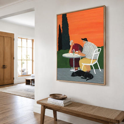 May include: A framed rectangular wall art print depicting two people sitting at a round white table outdoors against a vibrant orange sky. One person wears a dark red dress, the other a plaid shirt and yellow trousers. A black cat lies on the ground.