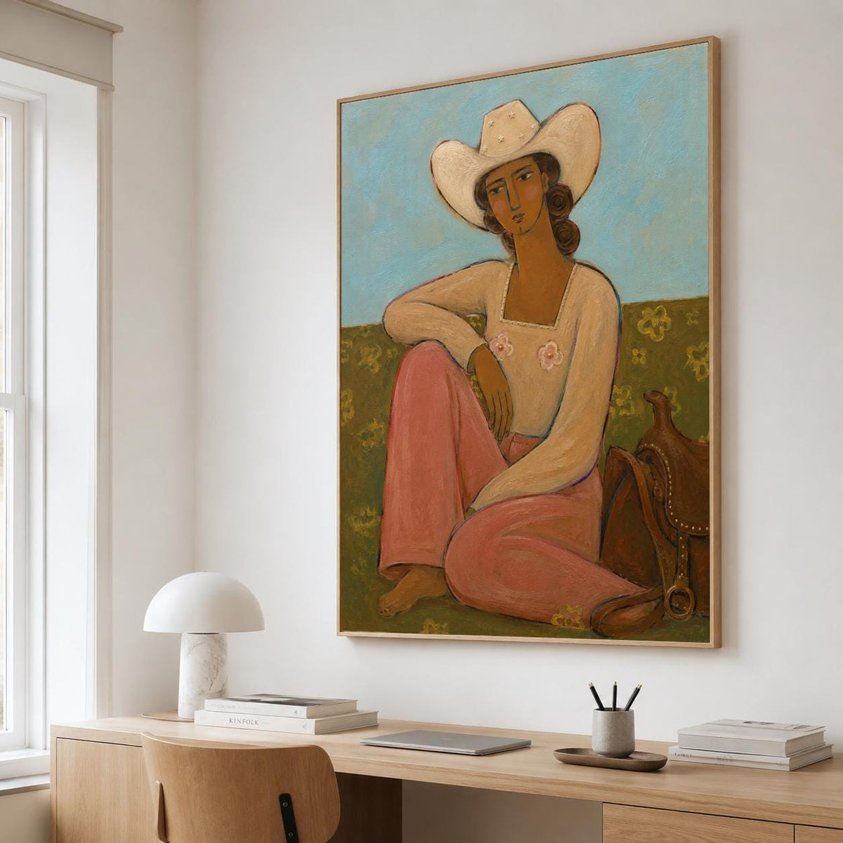 May include: A framed wall art print depicting a figure wearing a white cowboy hat and a long-sleeved cream top with two small pink flower accents. The figure wears pink trousers and sits on a green field against a light blue sky, next to a brown saddle.