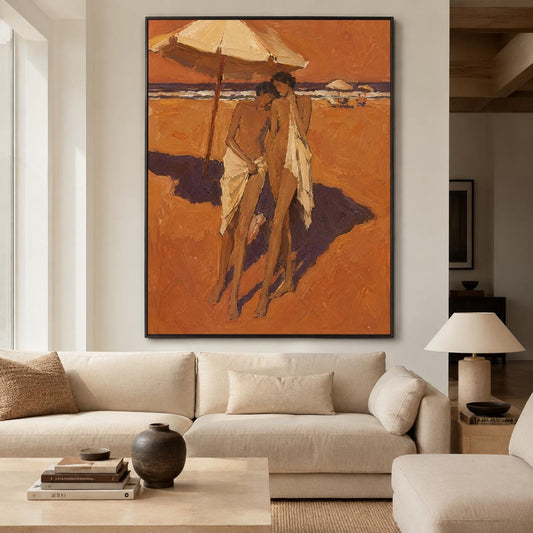 May include: A vertical oil painting in a black frame depicting two figures standing on a sandy beach under a large umbrella. The figures are draped in white fabric against a warm, orange-toned landscape with a dark blue ocean horizon line.